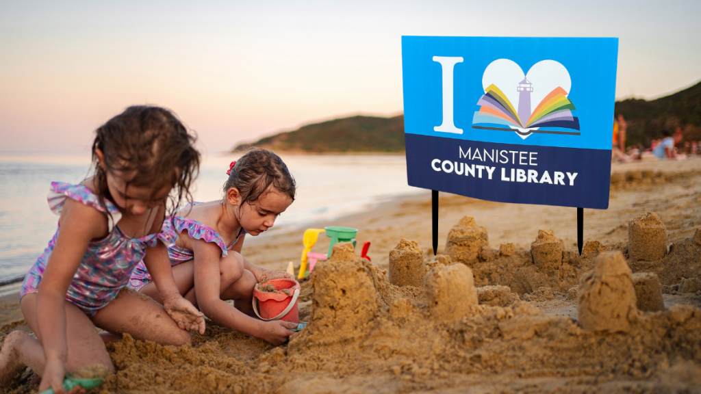 two kids building a sandcastle next to an I love Manistee County Library sign.